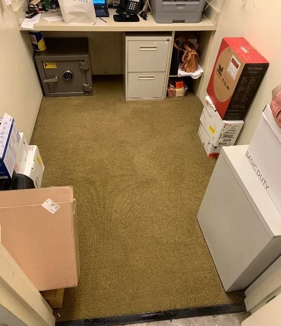 Small office storage room with tan carpet, a safe, filing cabinet, and boxes.