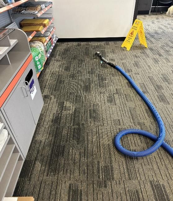 Wet carpet being cleaned with blue hose in a store; a caution sign is present.