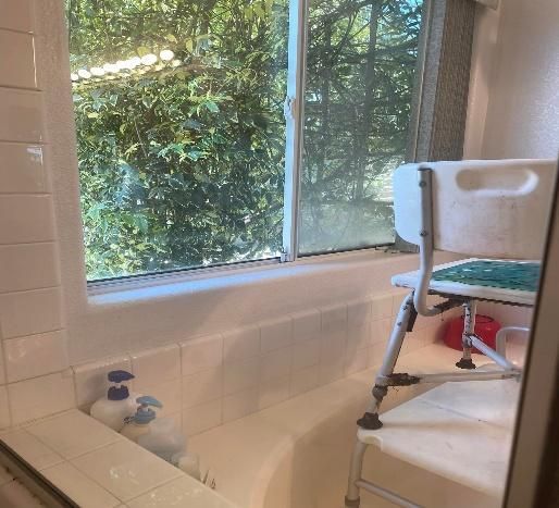 Bathroom with tub, window, and a white shower chair for accessibility.