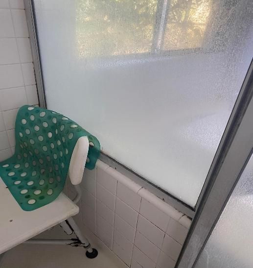 Shower stall with a green bath mat on a white shower chair, next to a frosted window.