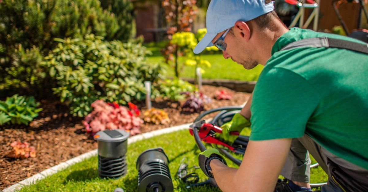 A man in a green shirt and cap uses a red tool on irrigation equipment. He is surrounded by garden plants and tools.