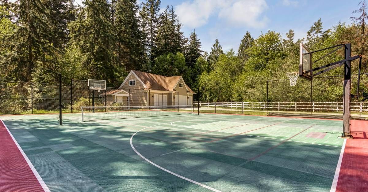 A green and red backyard basketball court with a house in the background surrounded by tall pine trees.