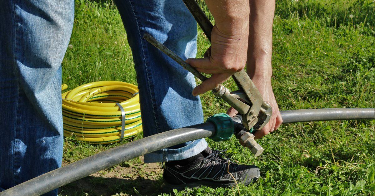  A person in jeans using a wrench to adjust a garden irrigation pipe, with a yellow garden hose coiled on green grass nearby.