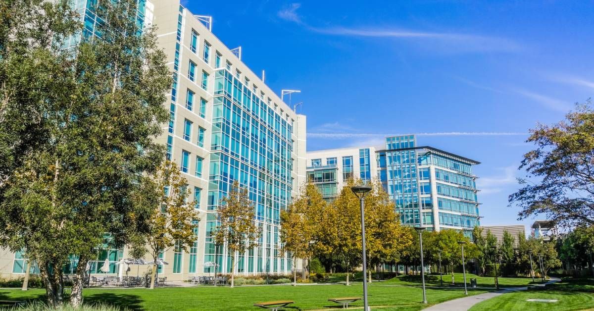 A landscaped urban park with green grass, trees, benches, and a curved path surrounded by glass office buildings.