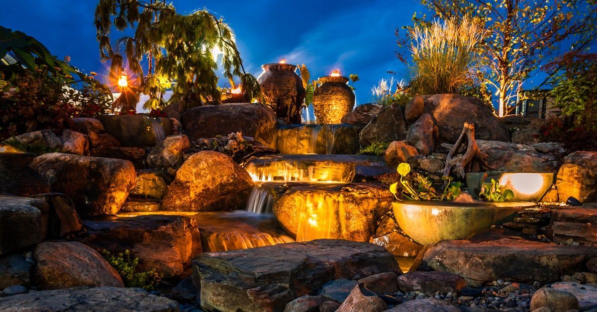 Landscape lights lighting up a boulder-waterfall area in a yard. It's dark outside and the lights are scattered in the rock.