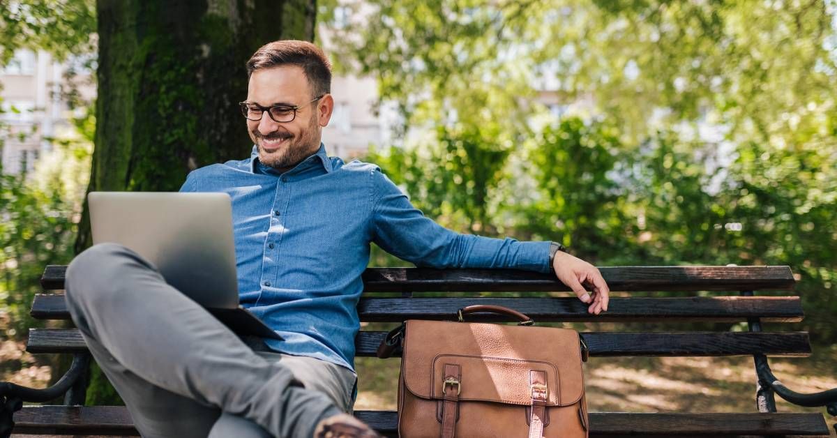 A man wearing glasses and a denim shirt sits on a wooden bench with a laptop on his legs and a leather bag beside him.