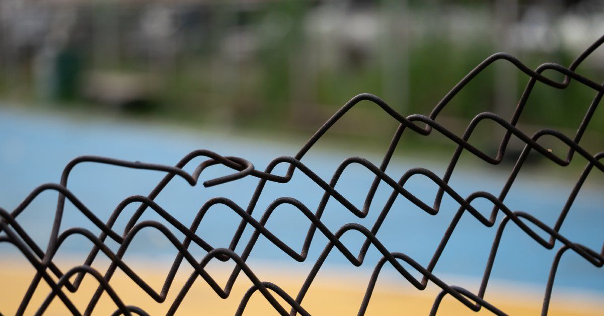 A damaged, bent chain-link fence at close range with a blue and yellow sports court blurred in the background.