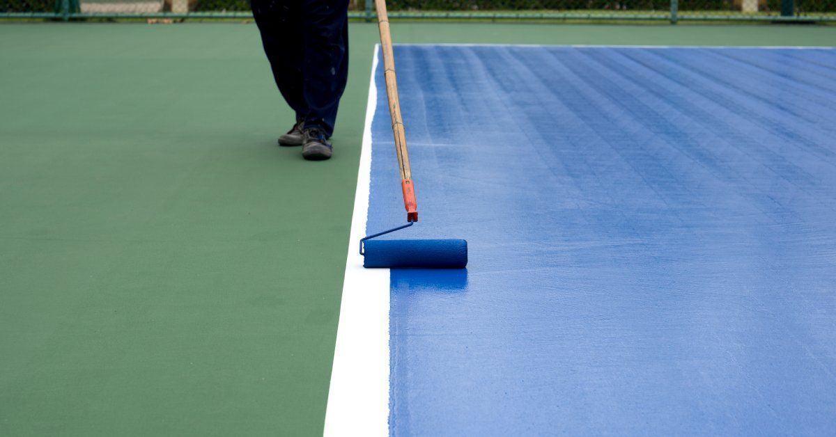 A person uses a paint roller on a long handle to apply blue paint over a tennis court, creating clear white boundaries.