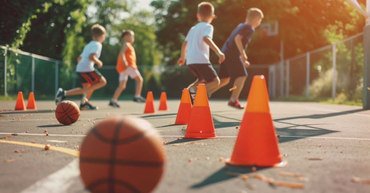 A group of four young boys playing outside on their home sport court. There are orange cones and two basketballs.
