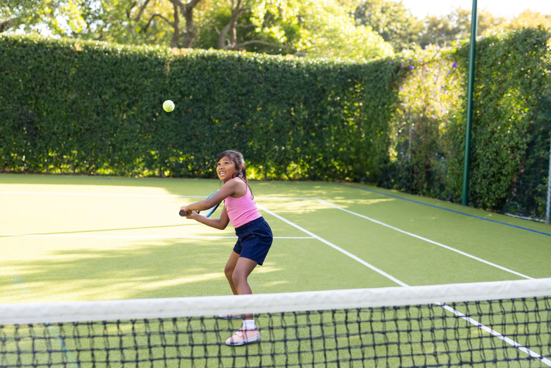 A young girl in a pink tank top playing tennis on her home sports court. The court is surrounded by 