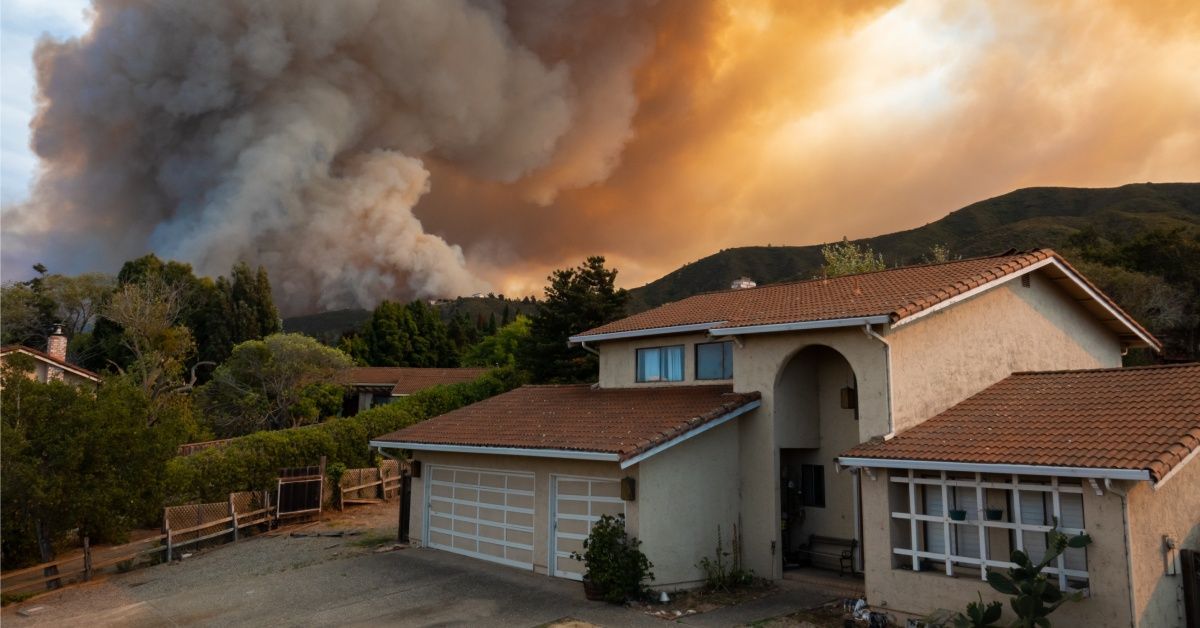 A California home on a small hill near trees. There's a wildfire taking over the trees and filling the air with smoke.
