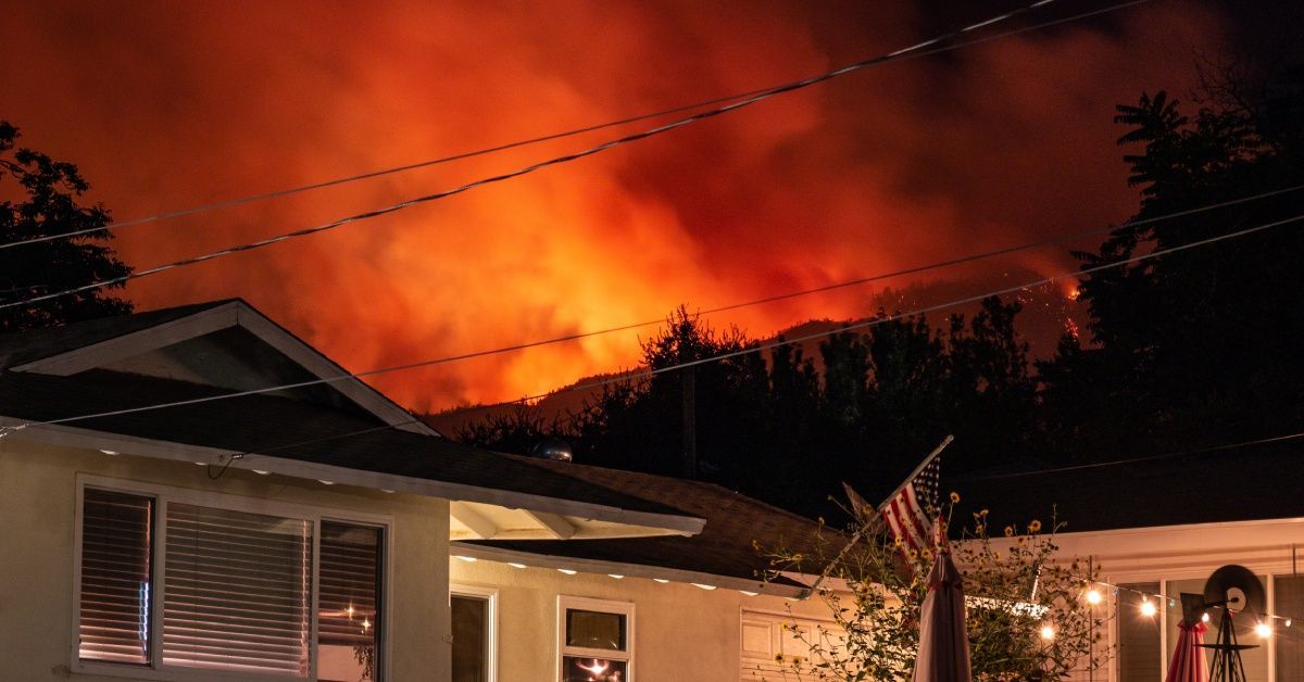 The top half of a residential home with an American flag waving. There is a massive wildfire raging behind the home.