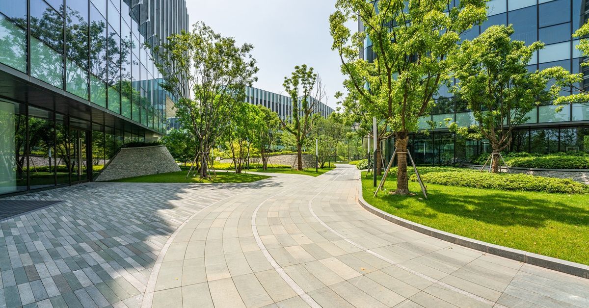 A modern office building area with a large paved walkway separating two buildings. The grass is green with small trees.