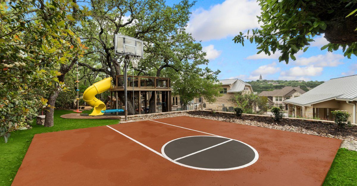 A backyard athletic court with a playground in the back. The court is brown with a basketball hoop at the end.