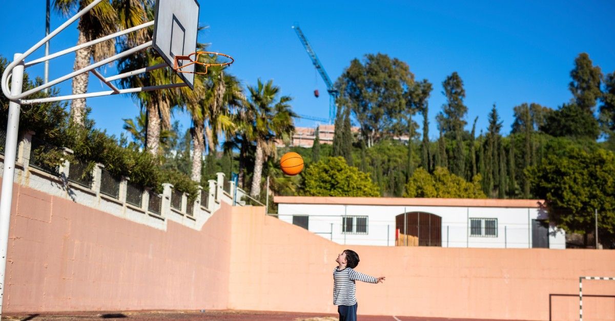 A small boy stands outside on the athletic court as he tosses a basketball into the air. It's sunny outside.