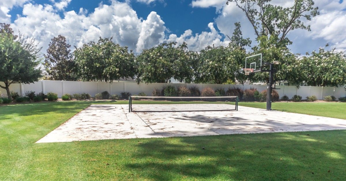 A residential pickleball court with a basketball net at one end of the court. The court is white with overgrown weeds.