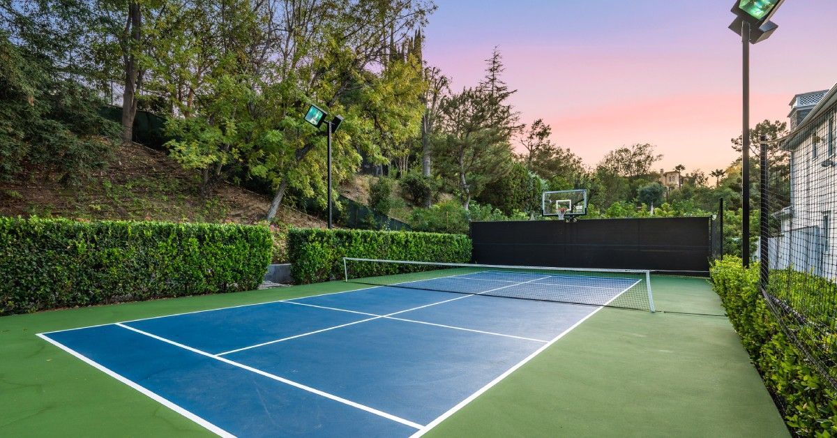 A residential tennis court with blue paint and white lines. Trees surround the court, and the sunset is light purple and pink.