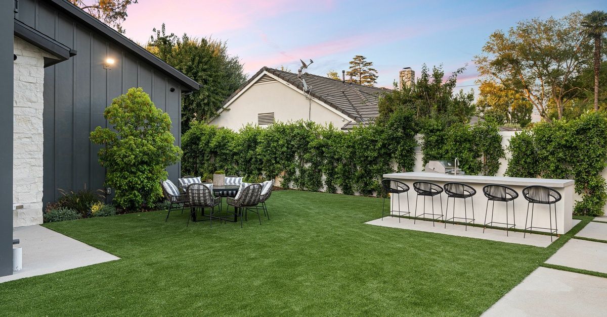 A beautiful backyard with synthetic turf, patio furniture, and a black house on the left. The sunset is blue and pink.
