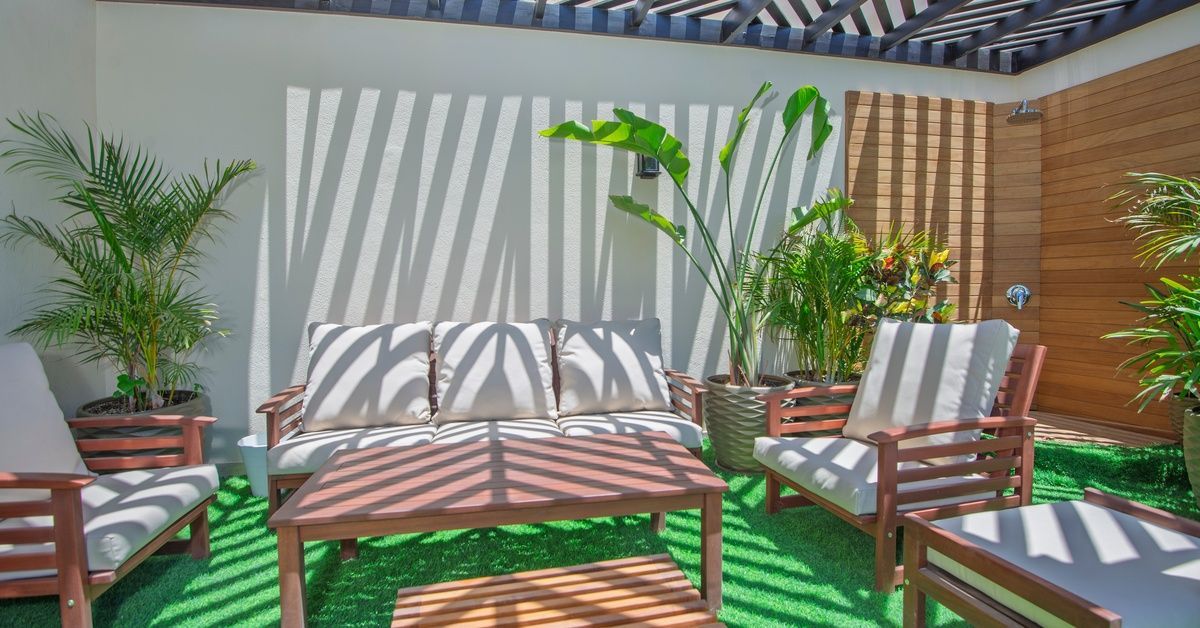 A sunny day in the backyard pergola with wood patio furniture, each one with white cushions. The ground is turf.