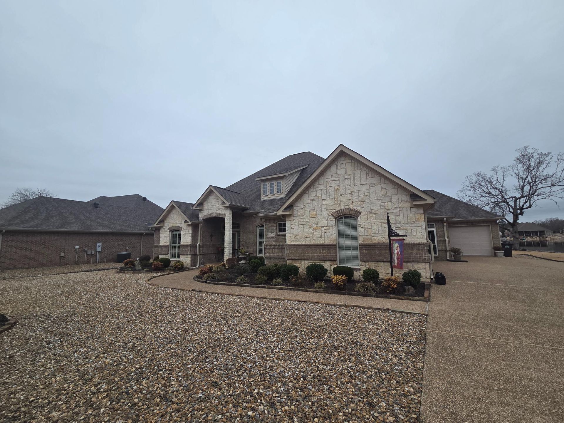 House with tan brick and brown roof on an overcast day. Driveway and yard covered in gravel.