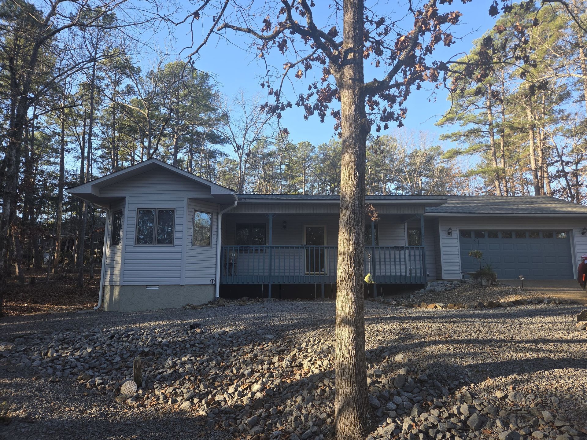 Gray house with porch, garage, and gravel yard, surrounded by trees under a blue sky.