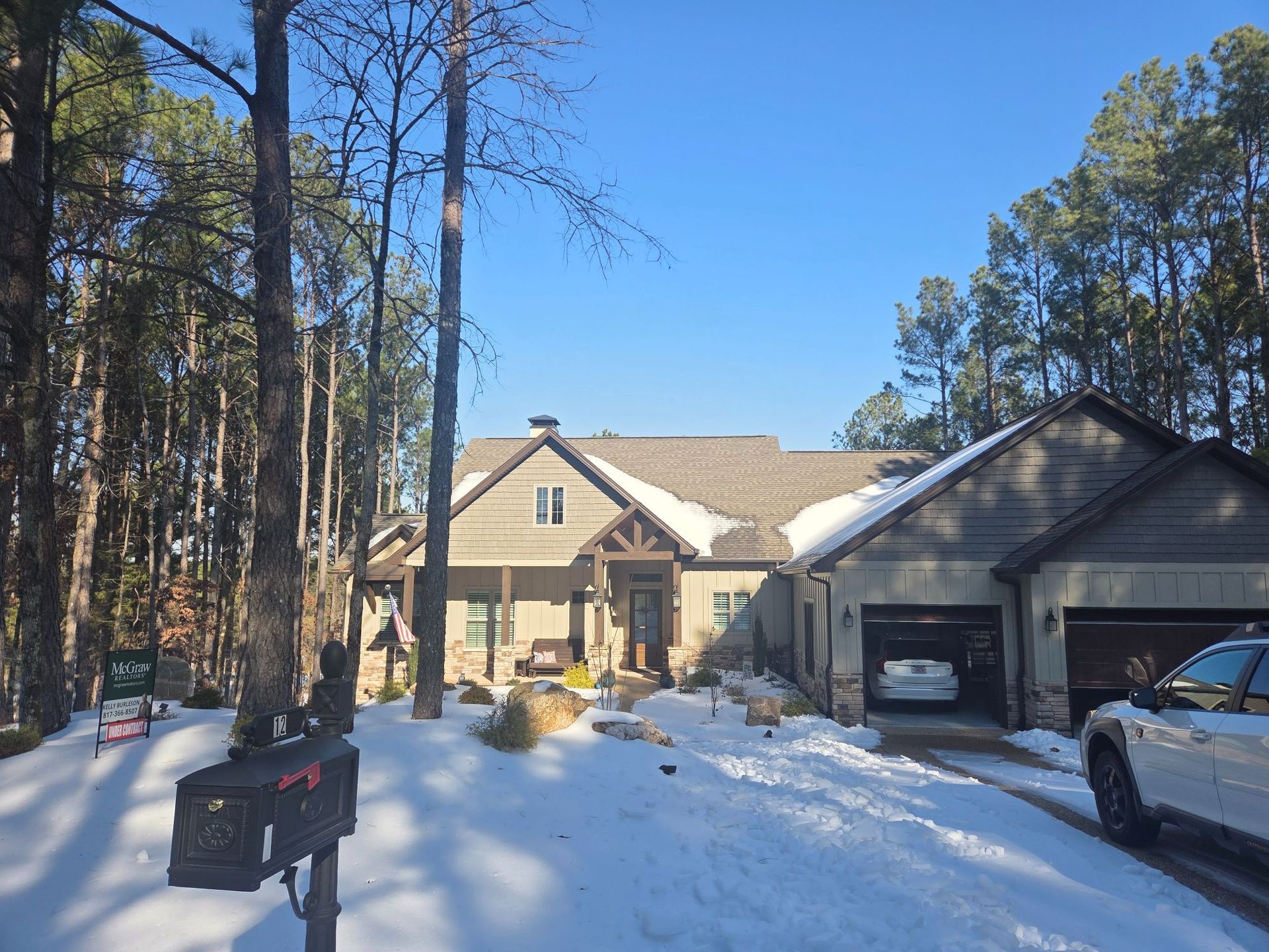 Snow-covered house with attached garage, mailbox in the foreground, and tall trees under a bright blue sky.