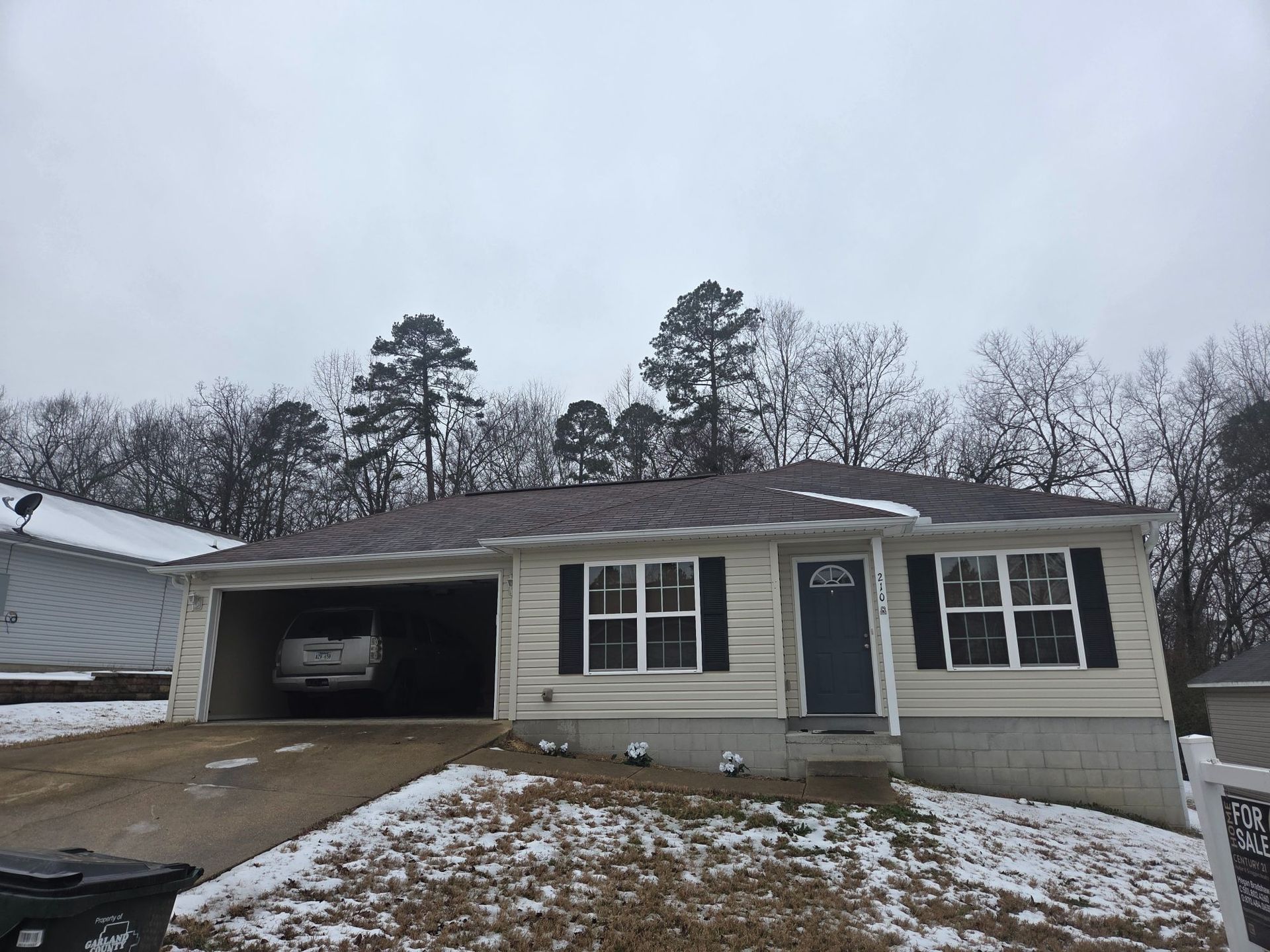 A single-story beige house with a driveway and garage, a car is visible inside. Snowy ground, gray sky.