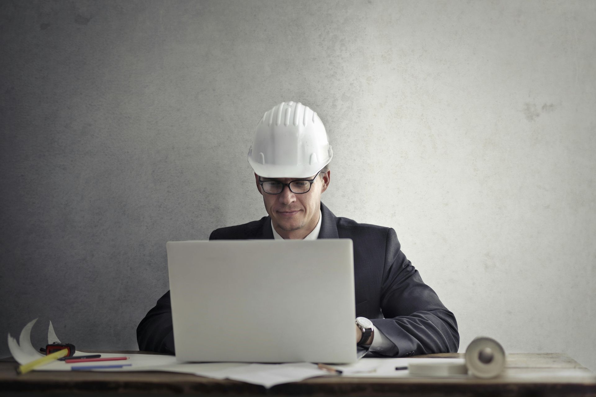 A man wearing a hard hat and glasses is sitting at a desk using a laptop computer.