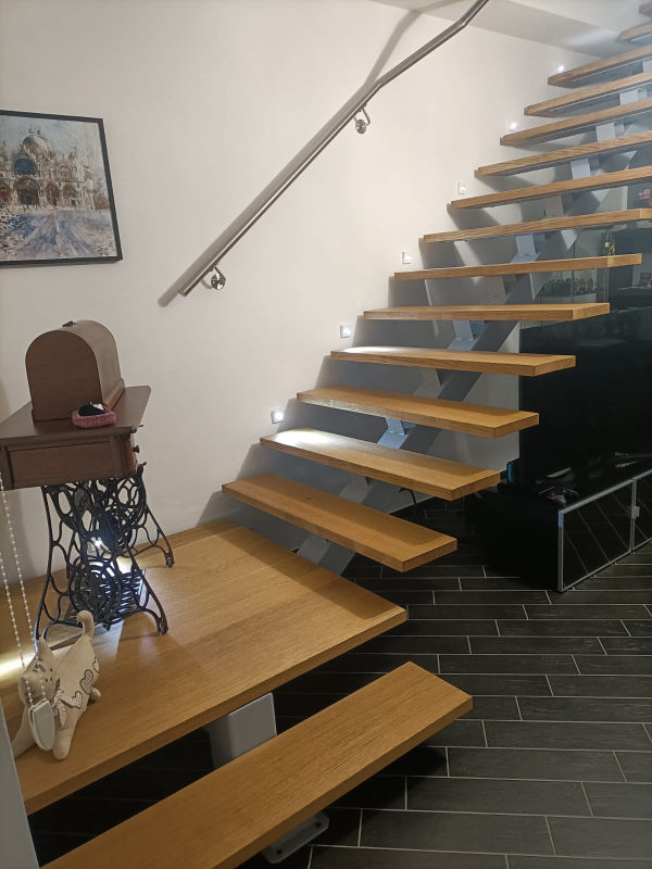 Scala moderna sospesa con gradini in legno chiaro, ringhiera in vetro e applique a parete sopra un tavolo da macchina da cucire vintage.