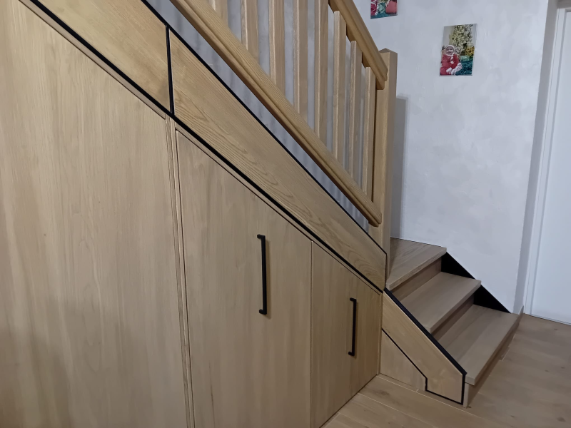 Armadietti sottoscala in legno chiaro con maniglie verticali nere, posizionati accanto a una scala in legno e a una parete bianca.