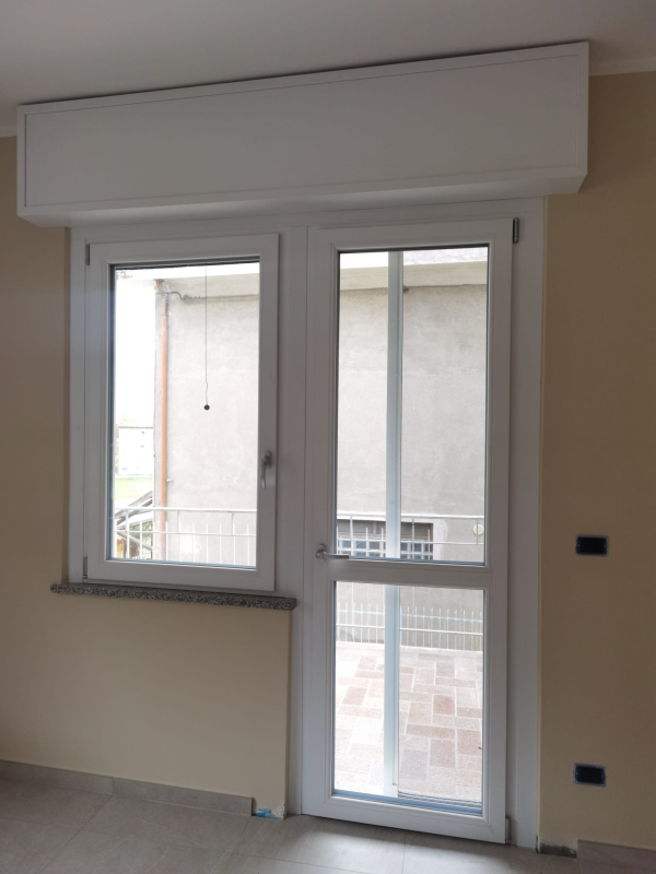 Una combinazione di finestra e porta con telaio bianco e cassonetto per tapparella integrato nella parte superiore, incastonata in una parete beige chiaro.