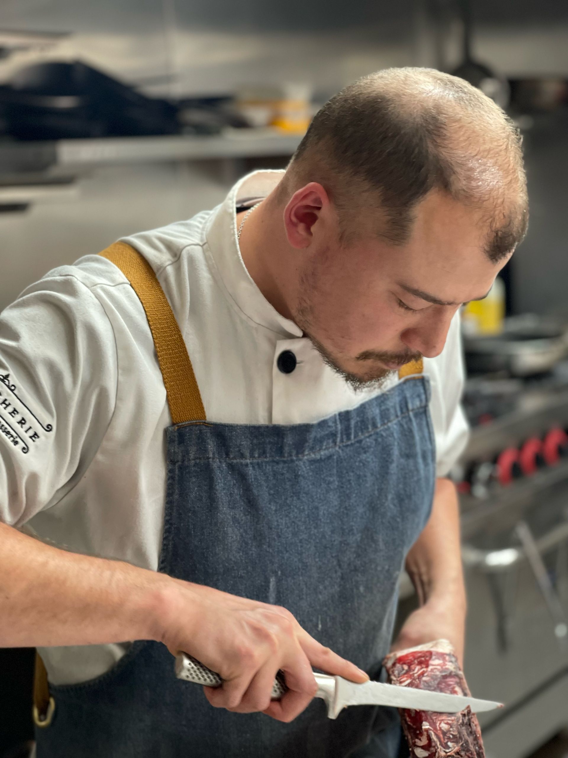 Chef in white coat and denim apron carefully cuts meat in a commercial kitchen.