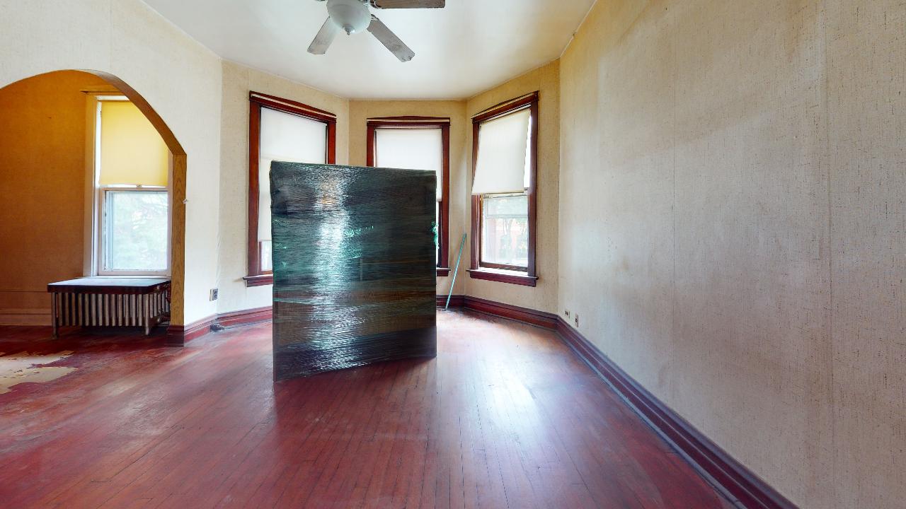 An empty living room with a ceiling fan and a large box wrapped in plastic.