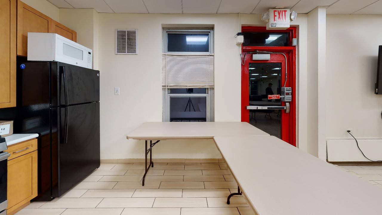 A kitchen with a table , refrigerator , stove and a red exit sign.