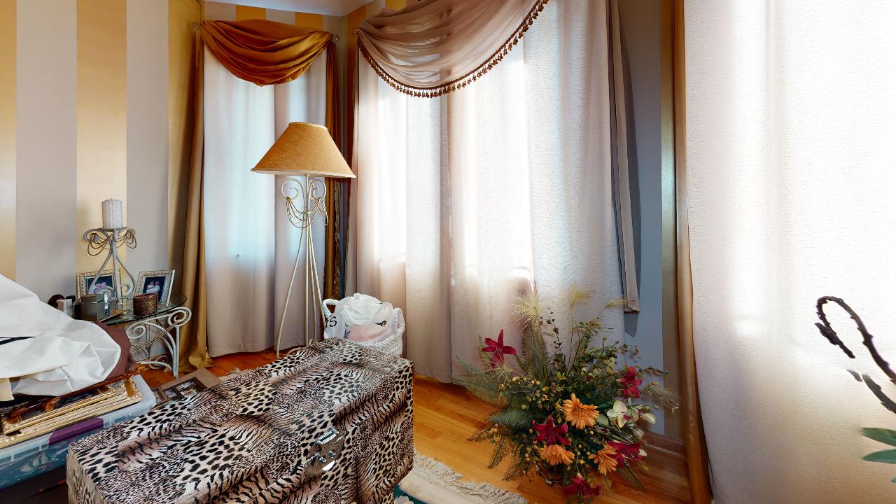 A bedroom with a bed , ottoman , lamp and a vase of flowers.