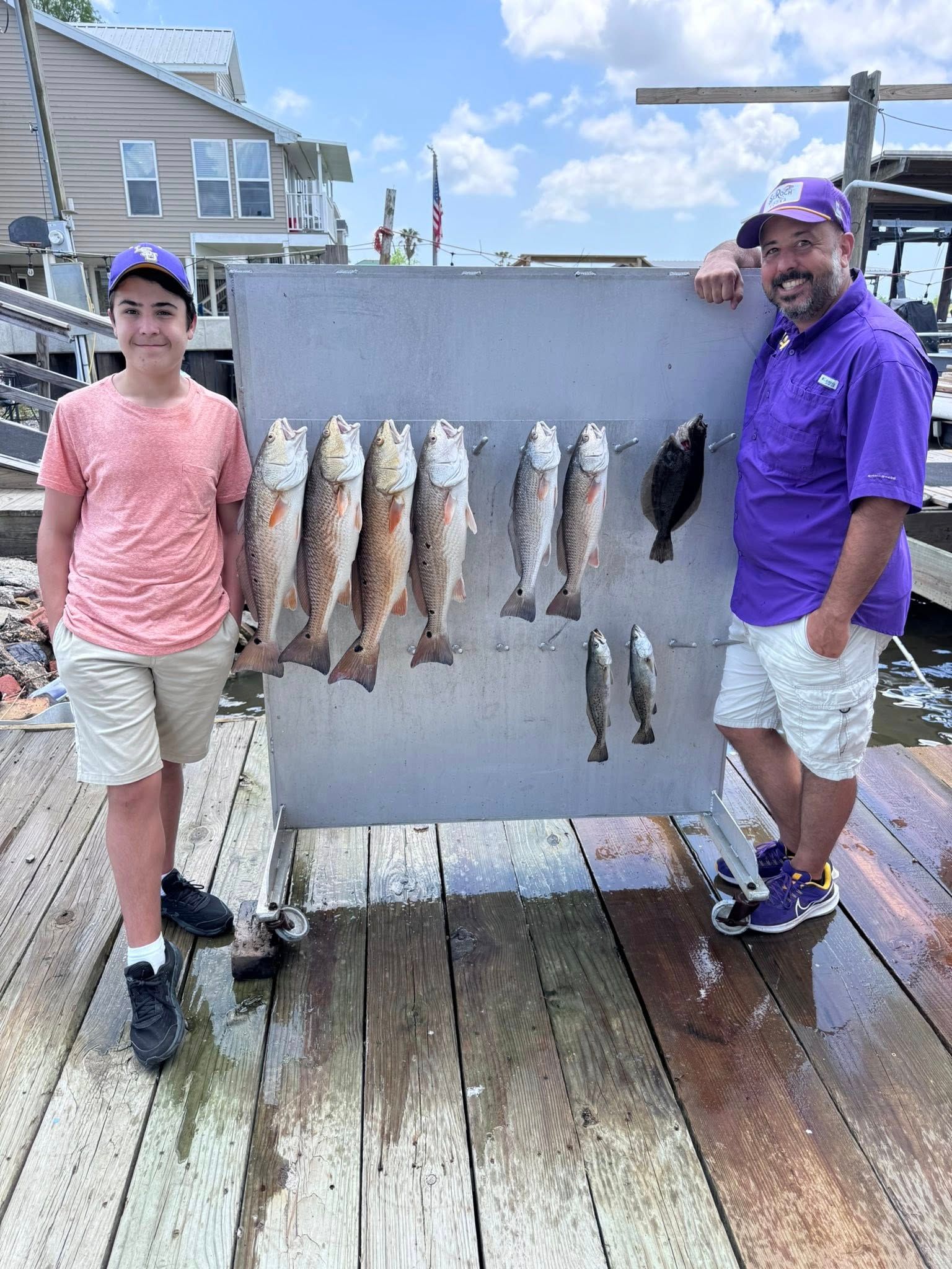 A man and a boy are standing on a dock holding a board with fish on it.