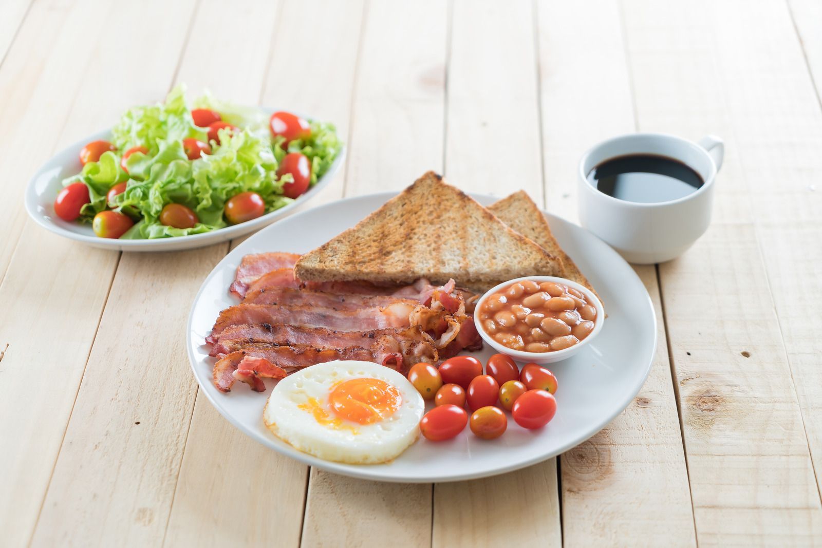 A plate of food with eggs , bacon , beans , toast and a cup of coffee on a wooden table.