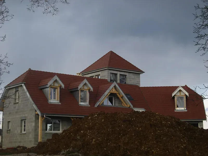 Maison inachevée avec toit de tuiles rouges, lucarnes et tour sous un ciel nuageux.
