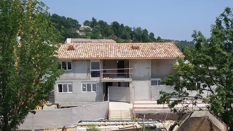 Bâtiment de deux étages en construction, avec toit de tuiles orange, fenêtres et balcon, entouré d'arbres et d'une colline.