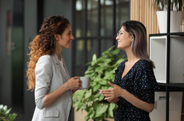 Two coworkers chatting with mugs in a modern office, smiling by a plant and shelving unit