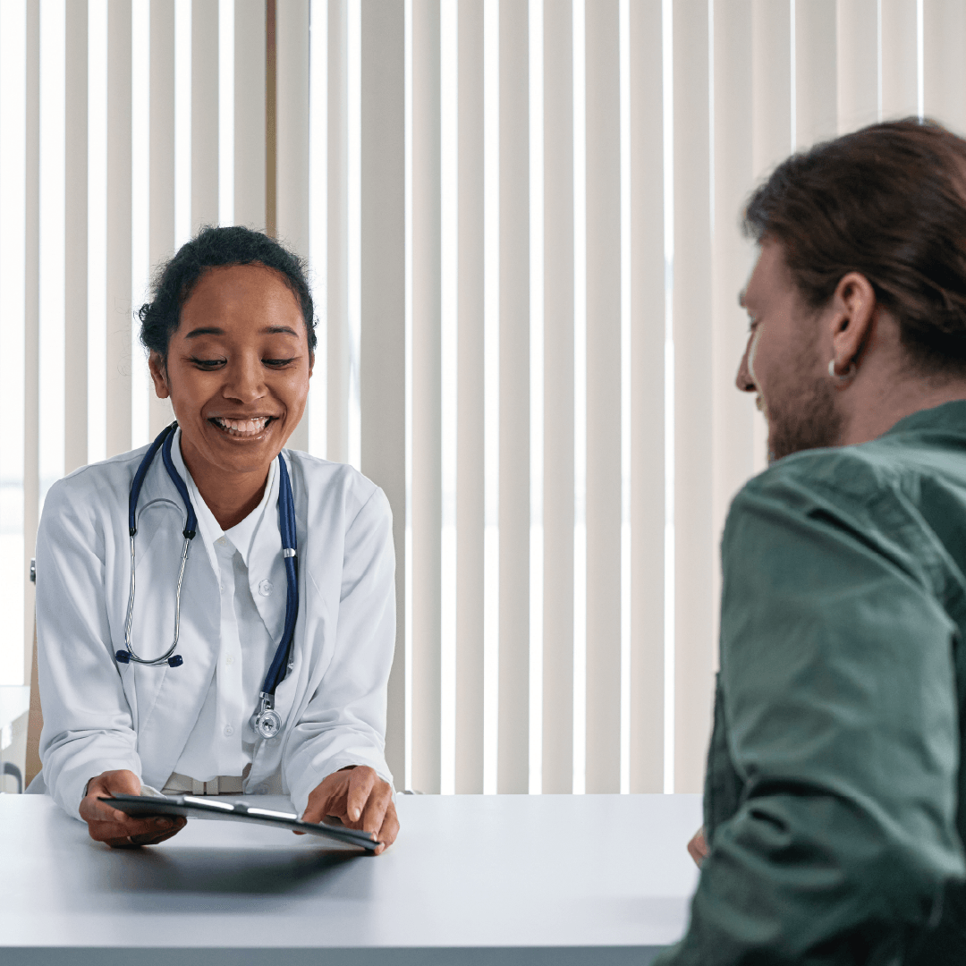 Médica sorridente conversa com paciente em consultório, ambos sentados frente a frente com ambiente claro ao fundo.
