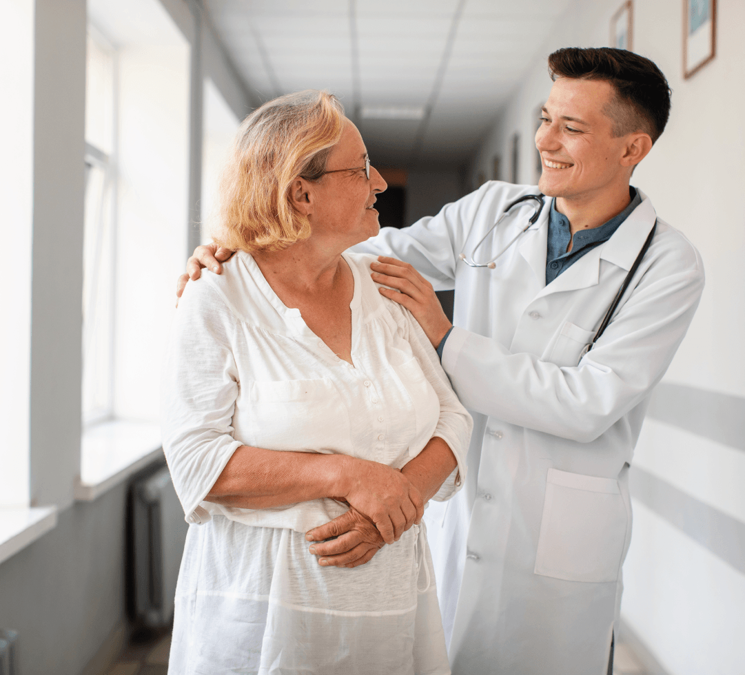 Médico sorridente conversa com paciente idosa em corredor de clínica, ambos com expressão amigável