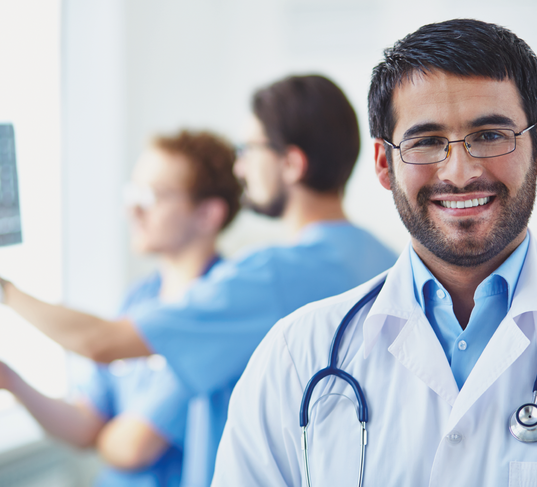 Corretor ou médico sorridente em destaque, com jaleco e estetoscópio, à frente de outros profissionais em ambiente clínico.