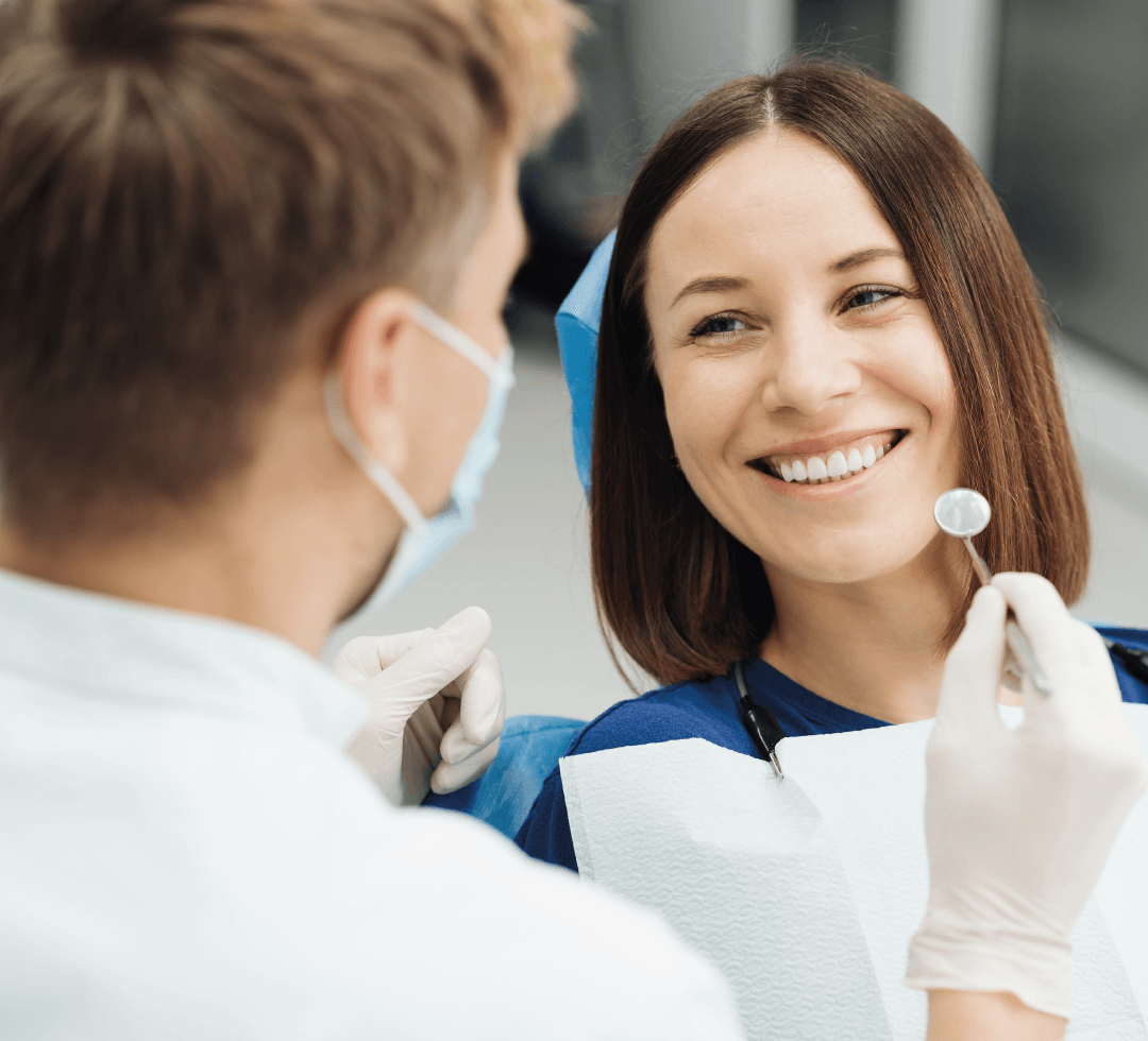 Mulher sorridente sendo atendida por um dentista, ambos usando avental e máscara, em um consultório odontológico.