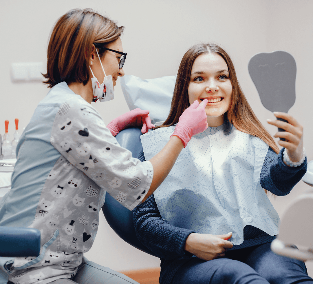 Jovem sorridente sentada na cadeira do dentista, segurando espelho enquanto profissional aponta para seus dentes.