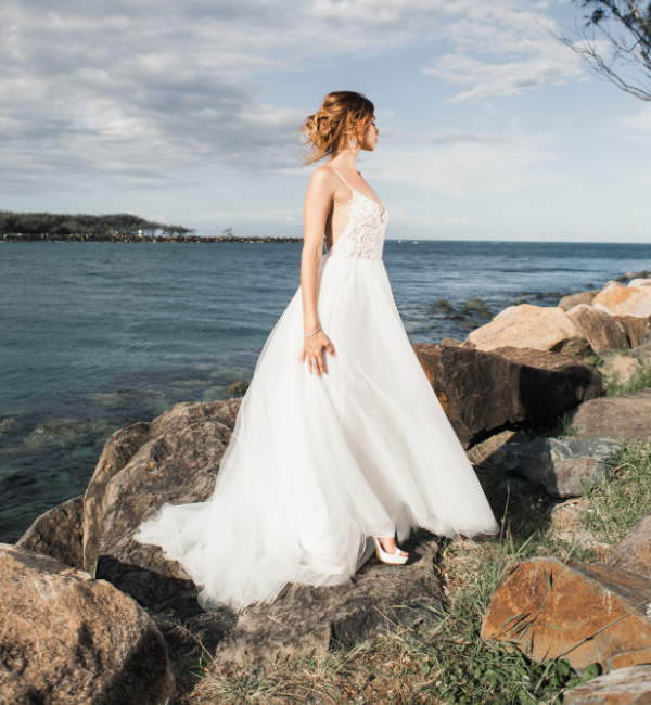 Bride in a white wedding dress standing on seaside rocks, looking toward the ocean.
