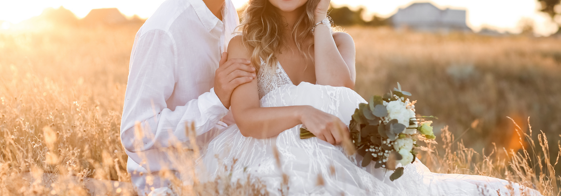 Bride and groom seated in a sunlit field, holding a bouquet amid warm golden light