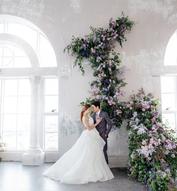 Bride and groom kissing in a bright white room beside a cascading purple and green floral arrangement