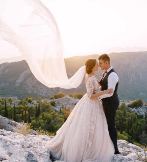 Bride and groom embracing on a rocky mountain overlook at sunset, with flowing veil and distant peaks