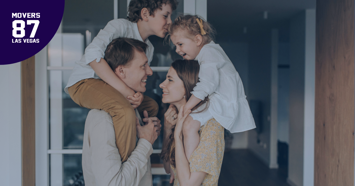 Family of four smiling, children on parents' shoulders, in front of a doorway.